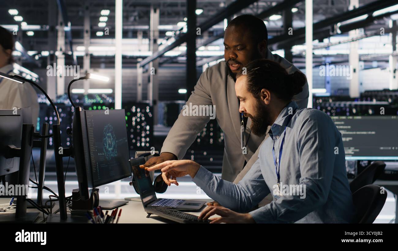 Data center engineers use laptop, collaborating on doing AI software updates. Server room teamworking colleagues doing brainstorming, optimizing infrastructure using artificial intelligence, camera A Stock Photo