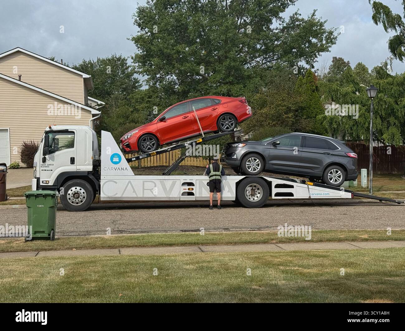 A flatbed Carvana tow truck loaded with a red sedan and a gray SUV on a suburban street with a man standing nearby - Smartphone Captured Stock Image