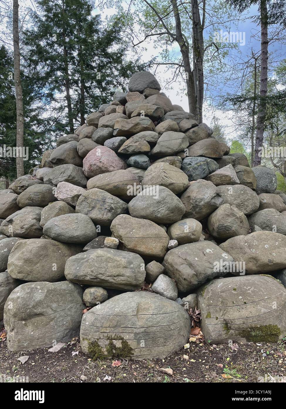 A large pyramid of stacked rocks in a forest clearing with trees and a cloudy sky in the background - Smartphone Captured Stock Image