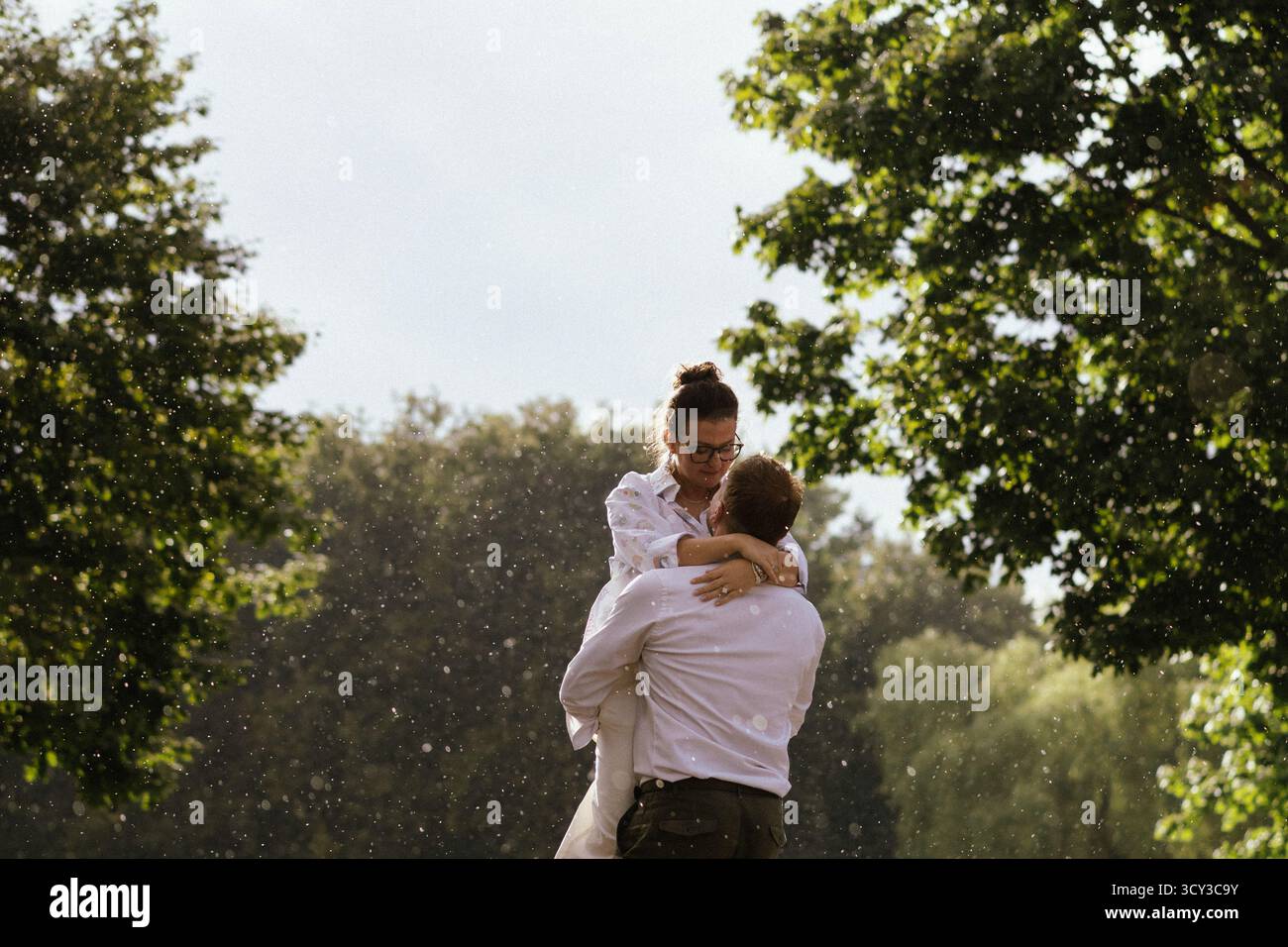 Romantic couple embracing and kissing in rain shower under green trees ...