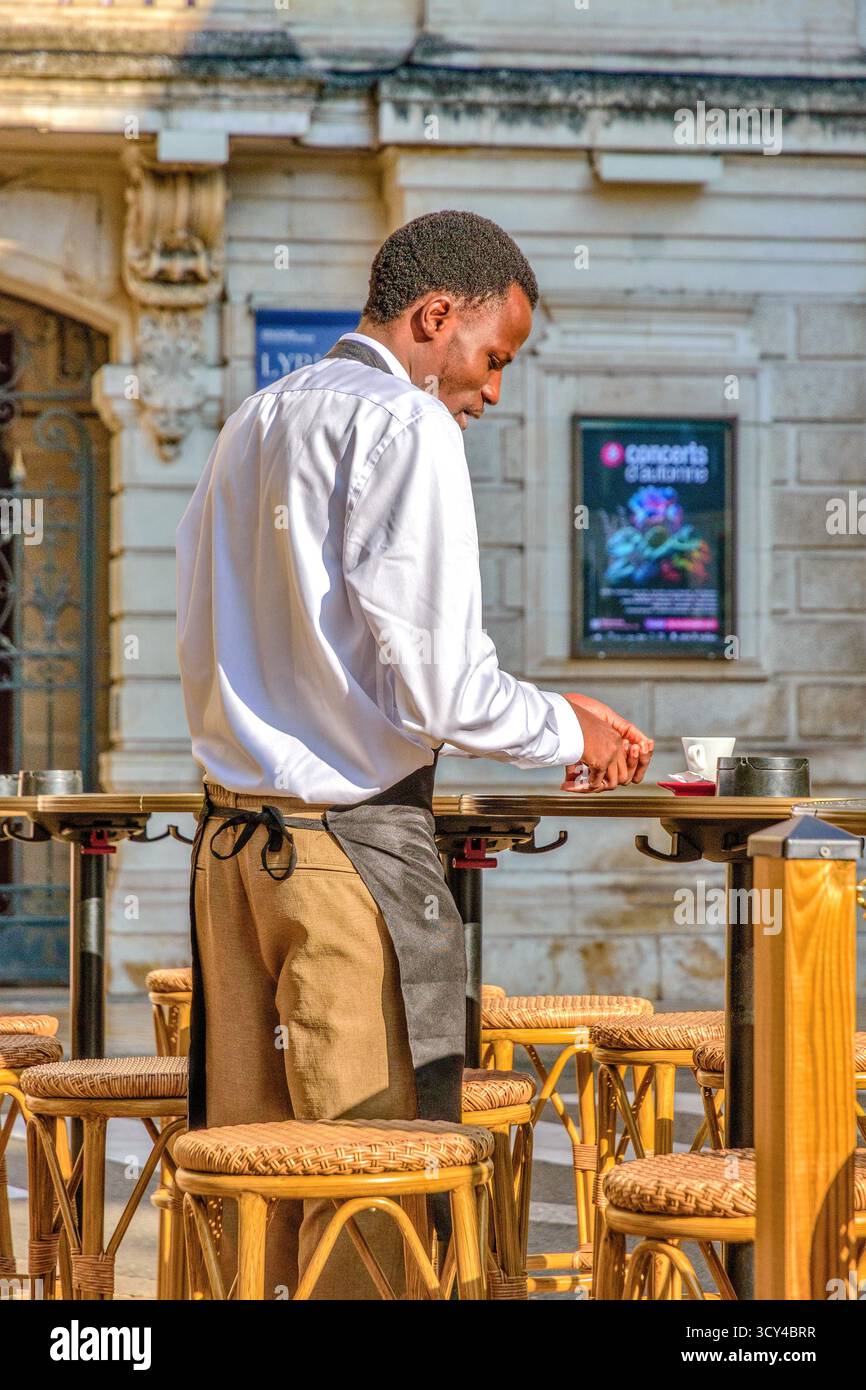 French-African restaurant employee clearing outdoor table of coffee cup - Tours, Indre-et-Loire (37), France. Stock Photo