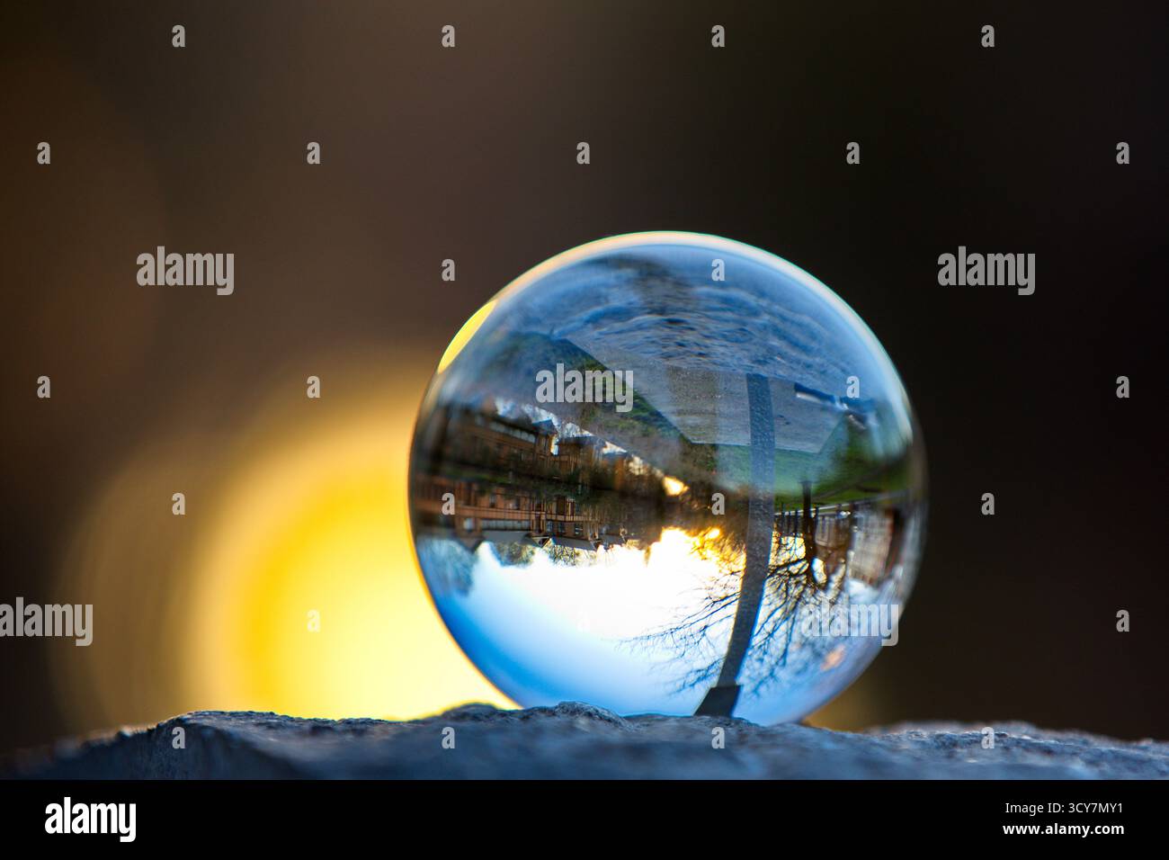 Crystal ball on a stone ledge reflecting an inverted riverside street and setting sun glow, captured with shallow depth of field at golden hour light. Stock Photo