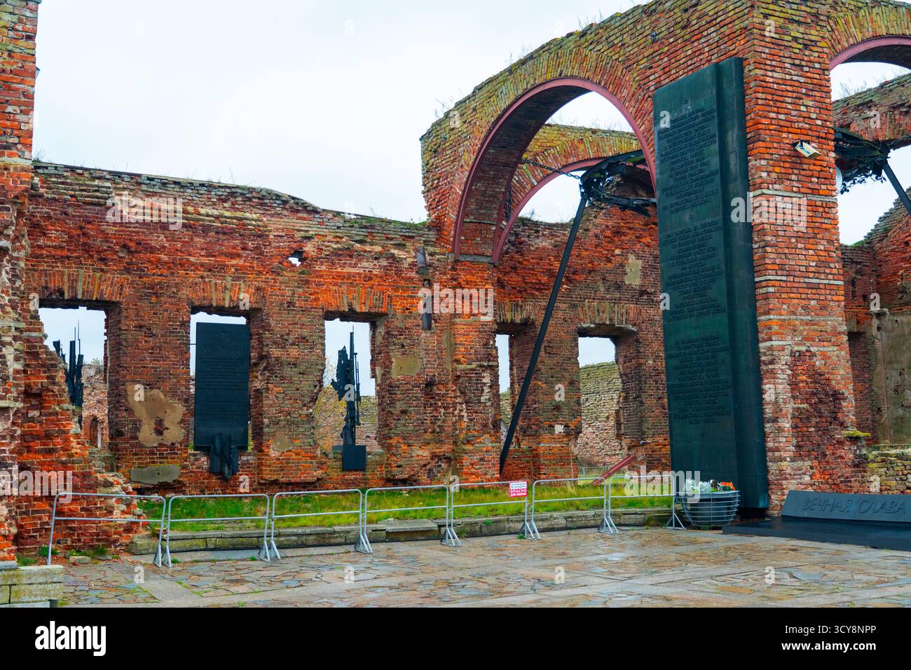 A black memorial stele standing amidst the red brick ruins and arches of a historic fortress under a cloudy sky Stock Photo
