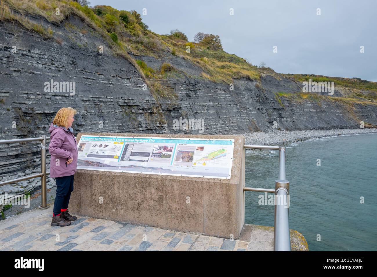 Woman reading the welcome to the Jurassic Coast World Heritage Site information board at Lyme Regis on the Dorset coast. Stock Photo