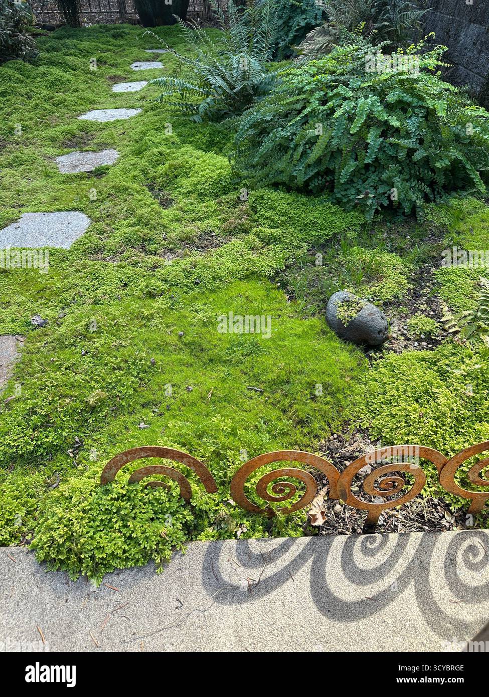 A shade garden with moss and ferns, a spiral metal border design, and slate stepping stones. Stock Photo