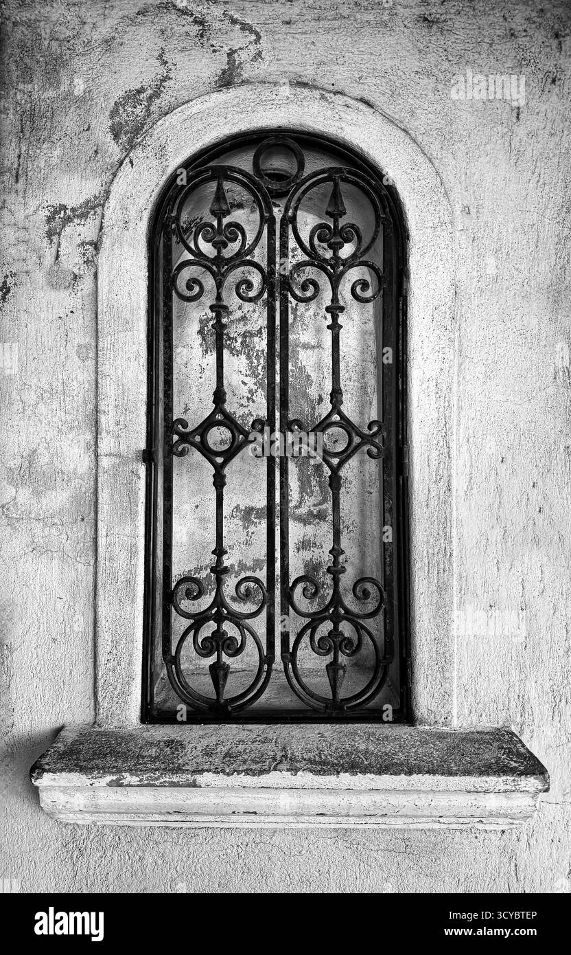 Black and white image of an ornate wrought iron grill on an arched window in an old stone wall - Smartphone Captured Stock Image