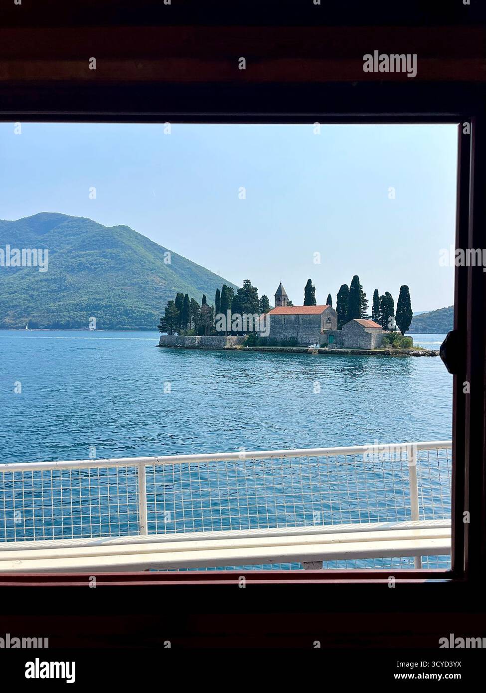 Peaceful island with a historic church and cypress trees viewed from a boat window, surrounded by calm blue sea and distant green mountains. - Smartphone Captured Stock Image