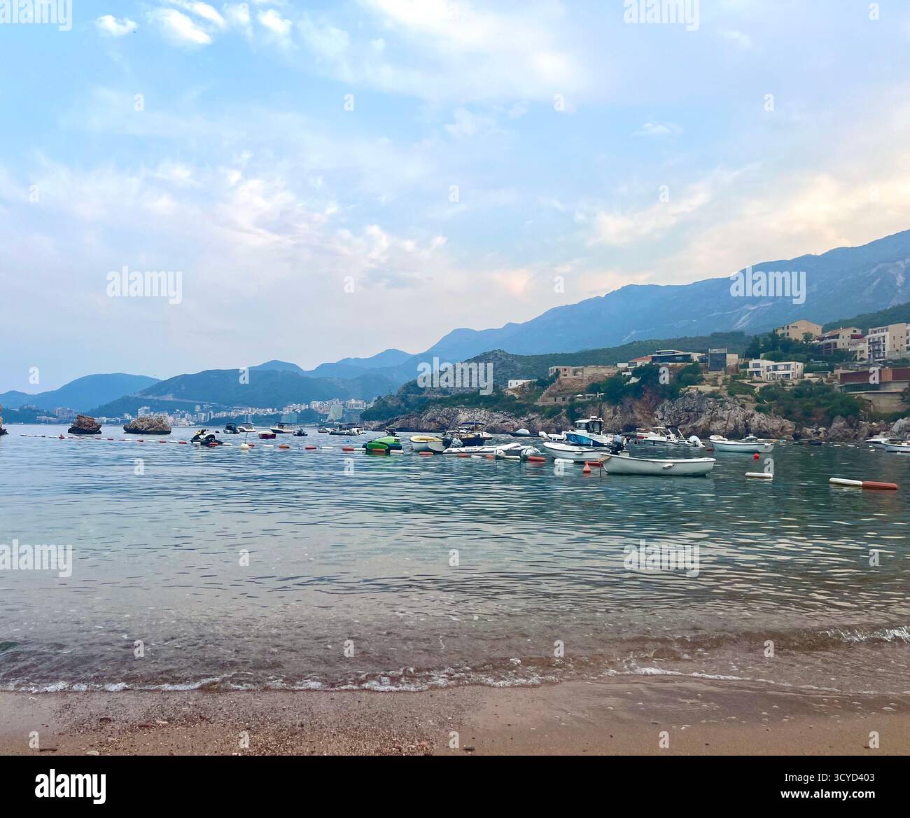 Peaceful coastal view with small boats anchored near the shore and mountains in the distance under a calm, cloudy sky. - Smartphone Captured Stock Image