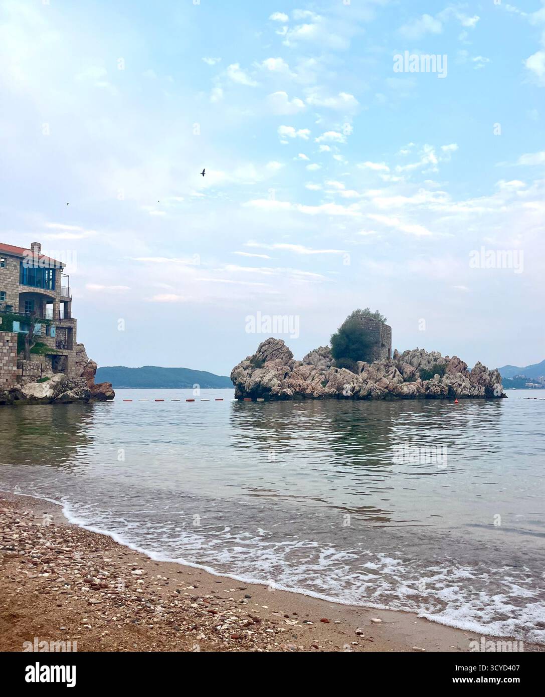 Seaside scene of the Adriatic Sea with a stone house, small rocky island, and gentle waves under a soft blue sky. Symbolizing coastal beauty. - Smartphone Captured Stock Image