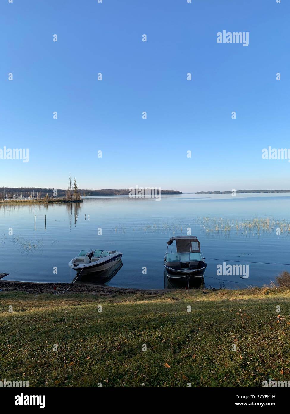 Serene autumn lake in Northern Canada with docked motorboats, grassy shore, reeds, and clear blue sky over distant hills. - Smartphone Captured Stock Image