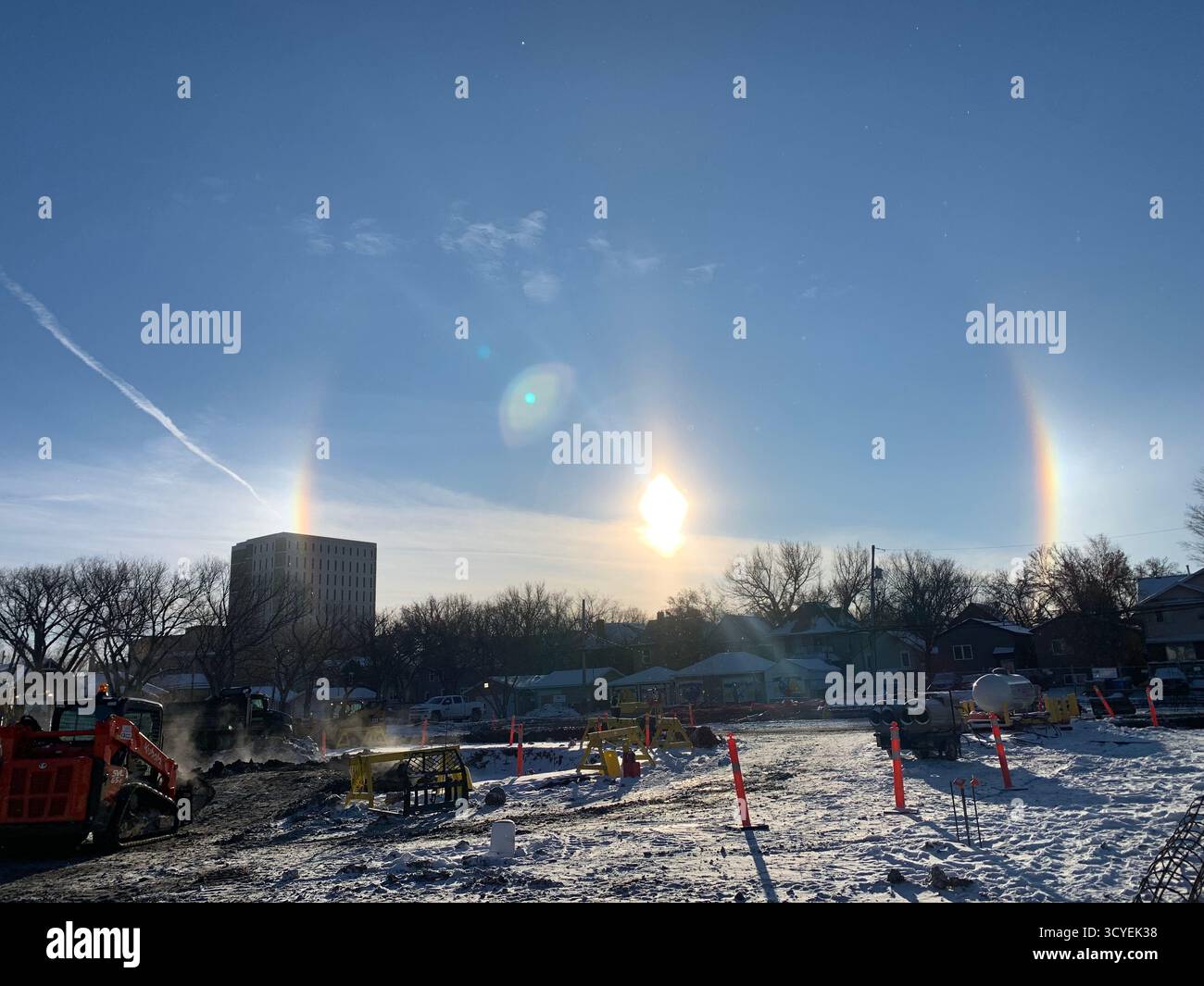 Sun halo and sun dogs over a snowy construction site with machinery and urban backdrop. - Smartphone Captured Stock Image