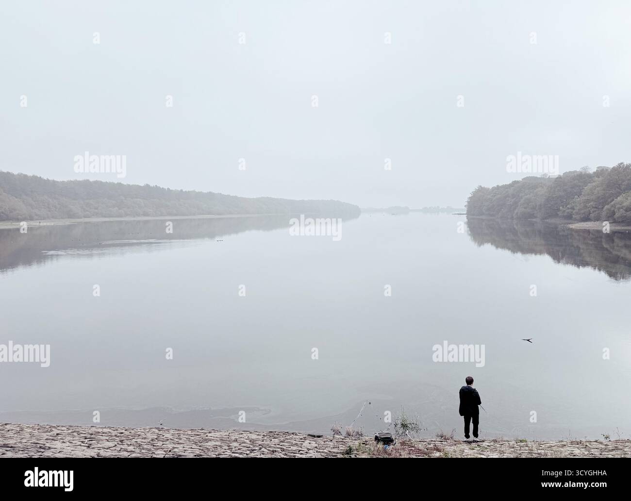 Person fishing in foreground and reflection of trees on a misty day at Rivington reservoir near Chorley in Lancashire - Smartphone Captured Stock Image