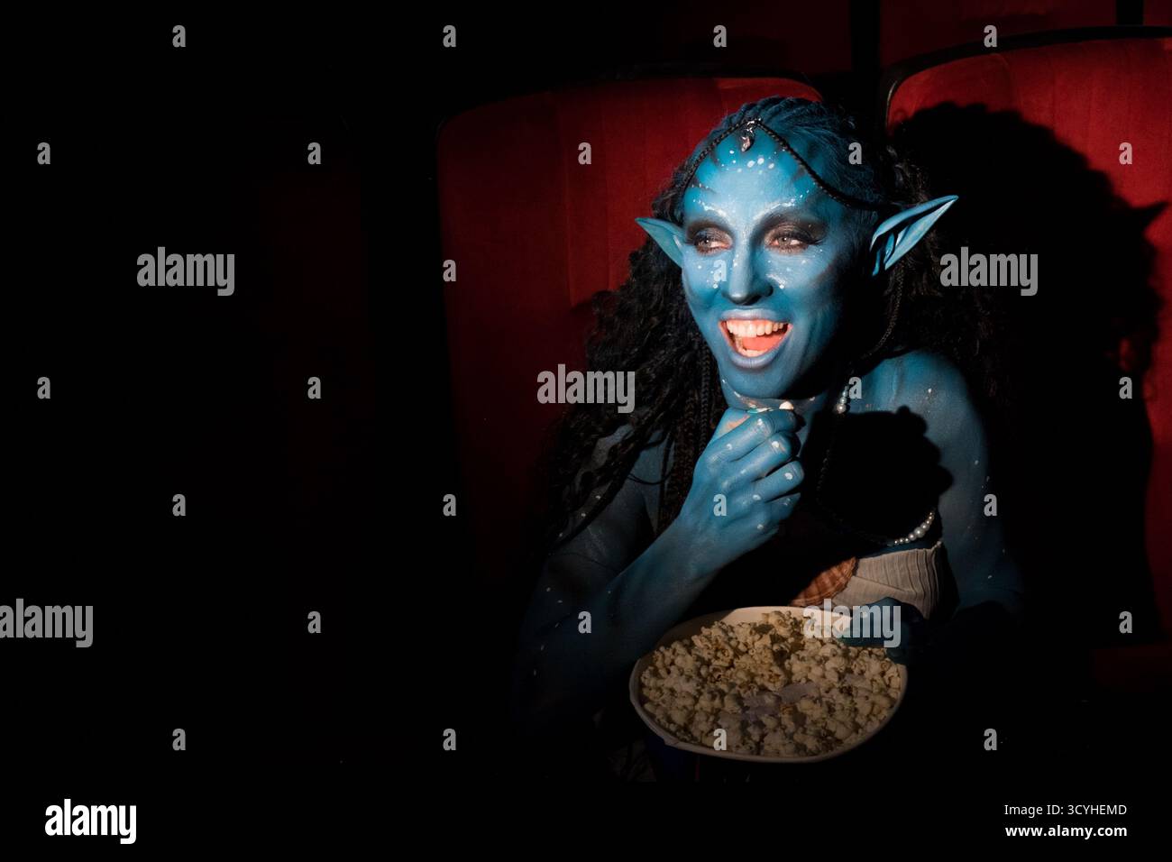 avatar, a girl in blue makeup is sitting in a movie theater and happily watching a movie with popcorn. The theater is dark. A private place for text. High quality photo Stock Photo