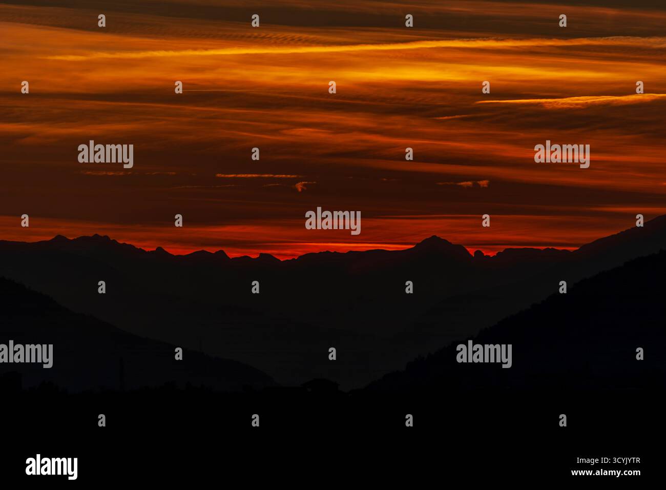 A stunning fiery sunset paints the mountain silhouettes with deep red and orange hues, creating a dramatic and peaceful alpine evening scene. Stock Photo