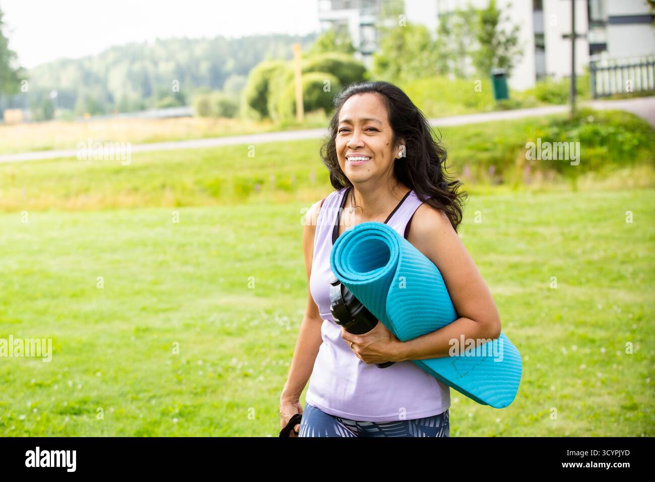Middle aged woman of color walking with yoga mat to park, embracing ...