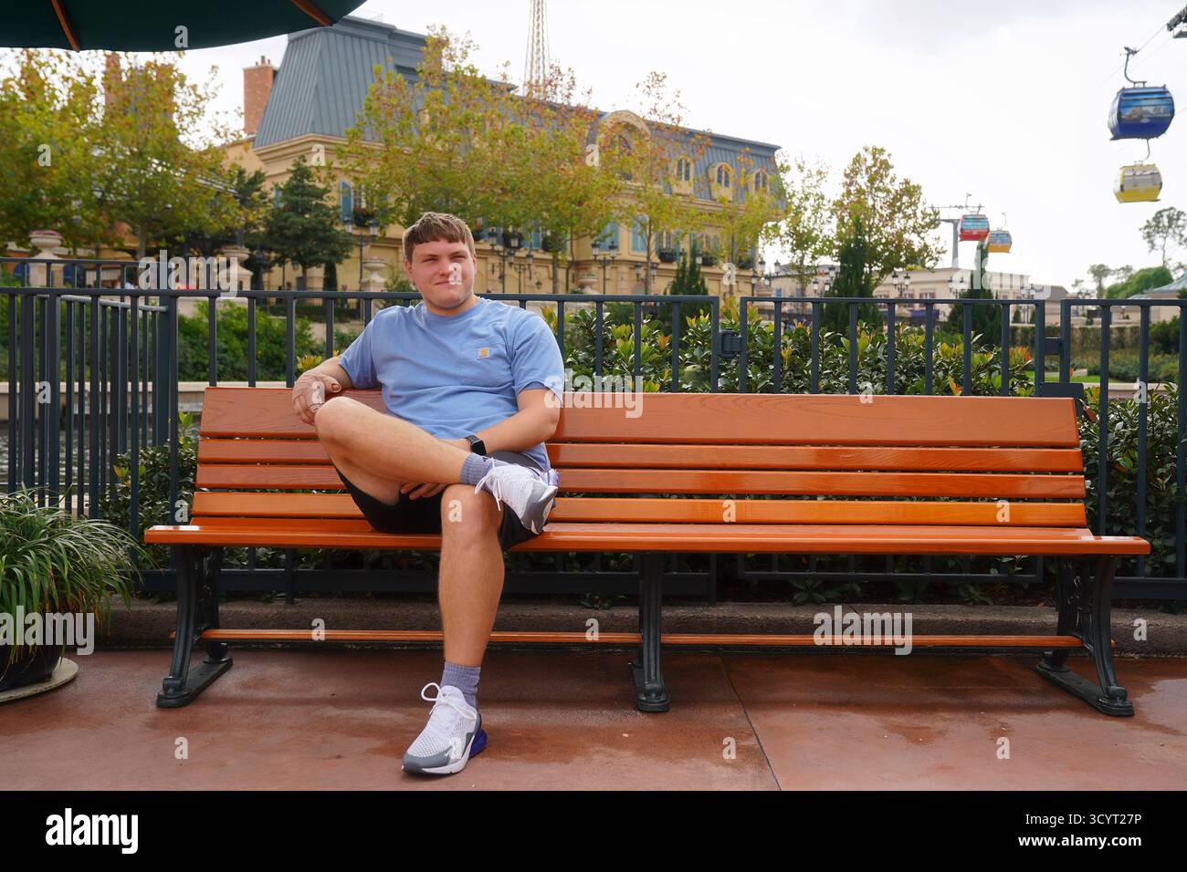 A young, caucasian man sits on a bench near the France Pavilion entrance at EPCOT, with the Disney Skyliner and Parisian-inspired buildings visible Stock Photo