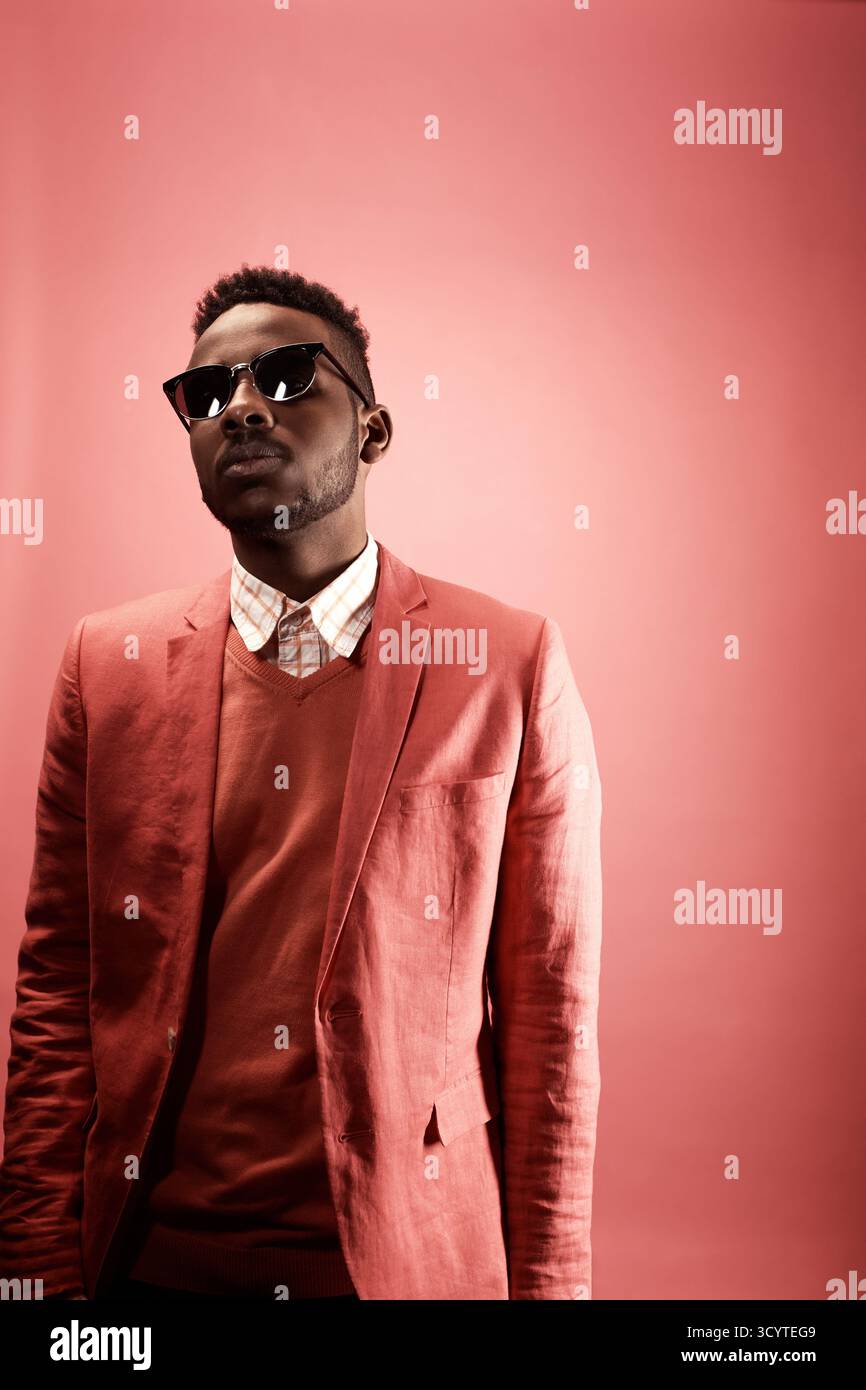 Studio portrait of young African American hipster man in stylish smart pink suit, shirt, knit jumper, sunglasses standing against plain red background, looking at camera Stock Photo