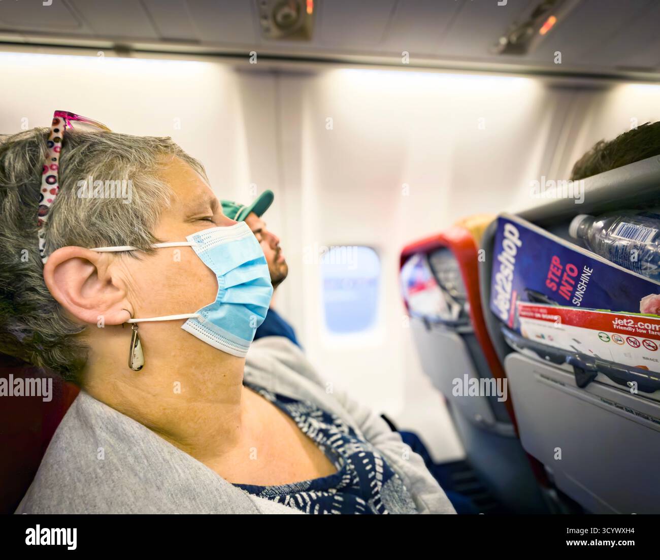 middle aged female Passenger wearing mask sleeping on a jet2 plane during daylight with sunlight coming through the window - Smartphone Captured Stock Image