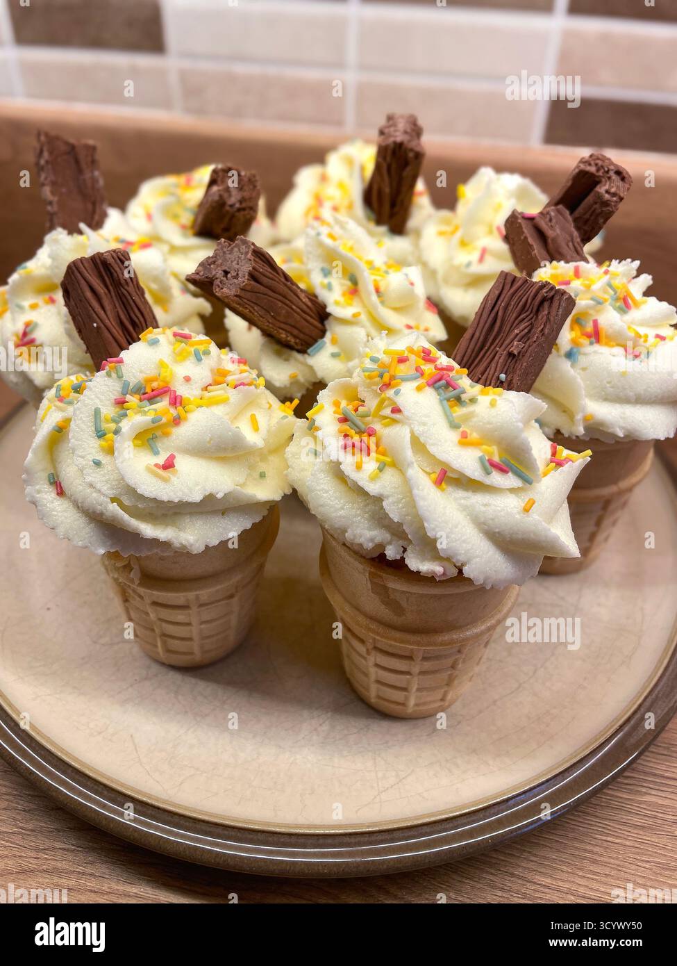 A plate of ice cream cone cupcakes topped with whipped cream, chocolate flakes, and colourful sprinkles - Smartphone Captured Stock Image