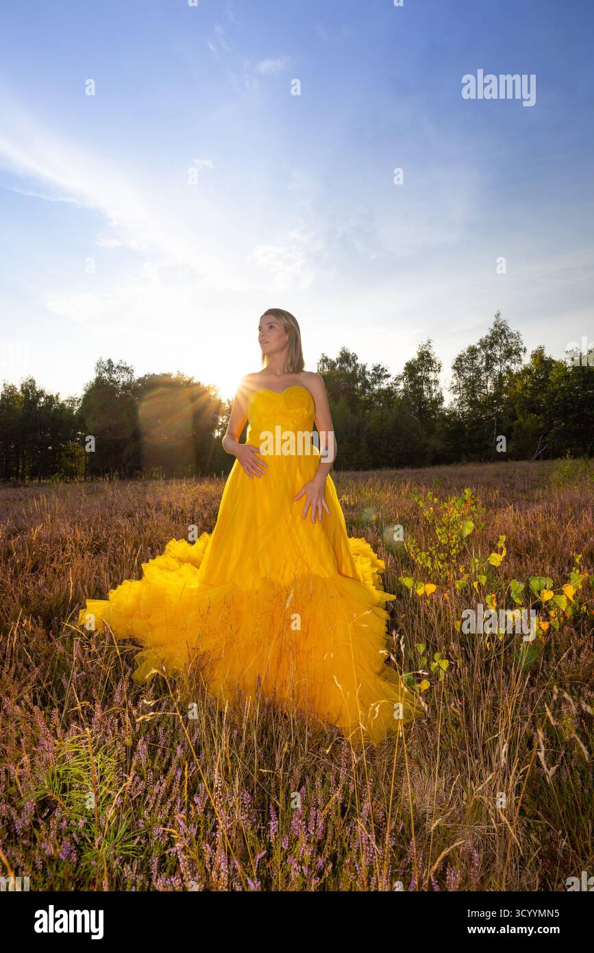 A woman elegantly poses in a stunning yellow gown amidst a blooming field as the sun sets beautifully Stock Photo