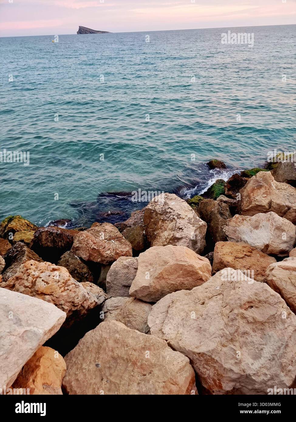 Beautiful seascape with turquoise waves, rocky coast, and a small island under soft pink sunset sky — peaceful natural scenery. - Smartphone Captured Stock Image