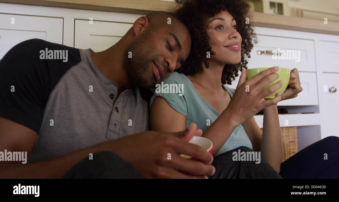 Leaning couple in raglan, teal shirts sitting on kitchen floor by cabinets, with green white mugs Stock Photo