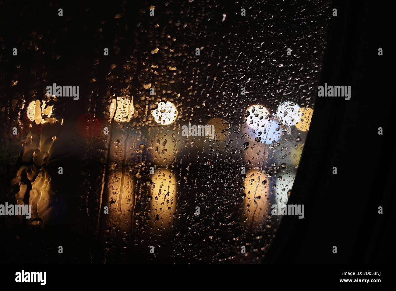 Raindrops are rolling down on a car window with blurred, bokeh city lights in the background at night Stock Photo