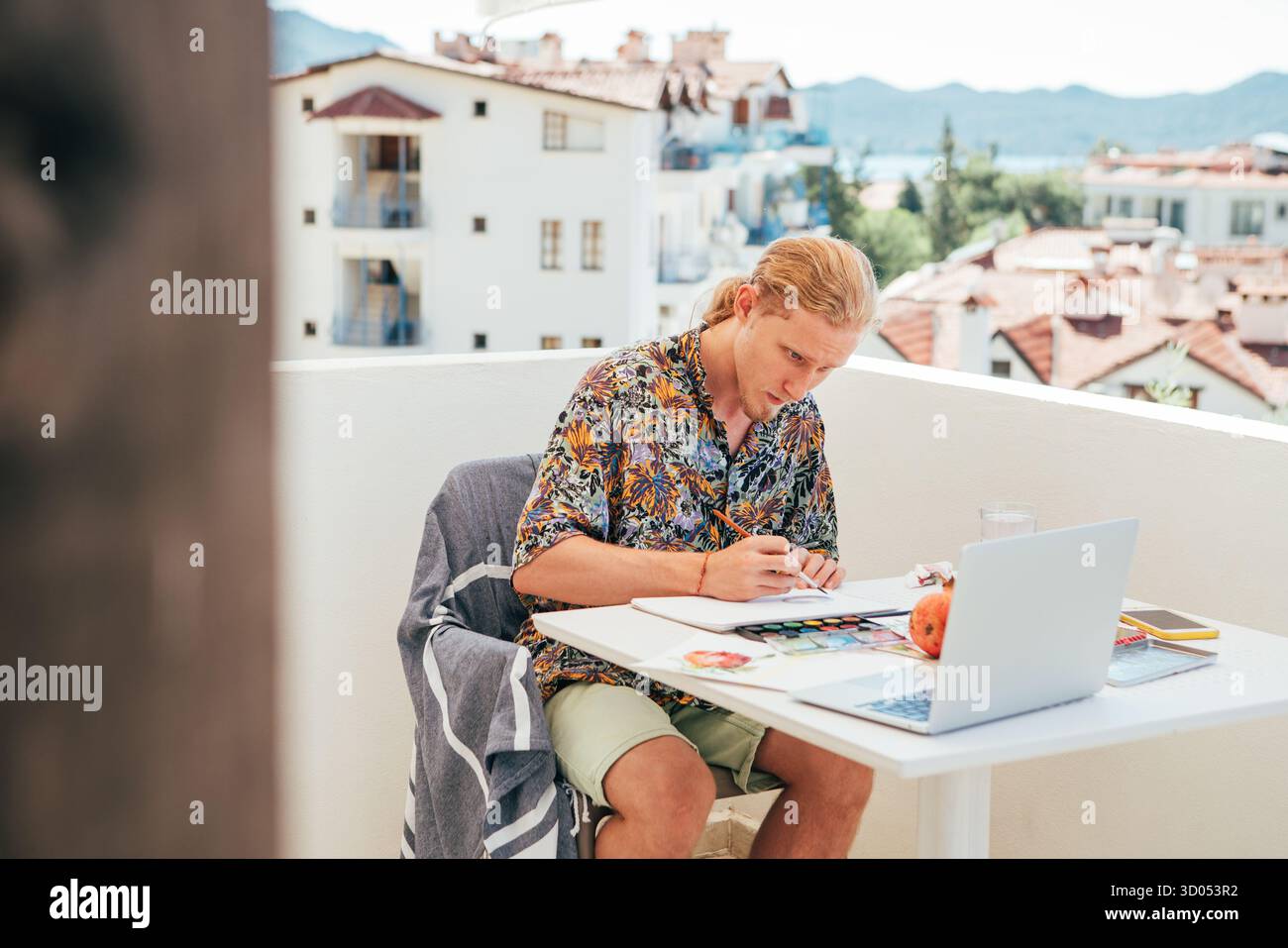 Teenager boy painter sitting on balcony creating Watercolor artwork during seaside summer vacation. He enjoying creative process and sunny coastal atm Stock Photo
