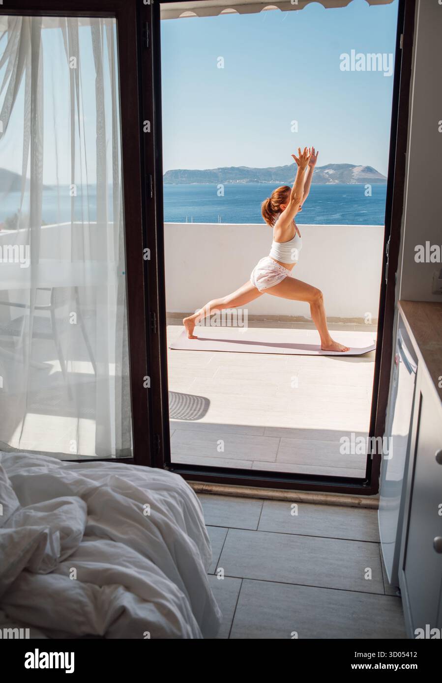 Middle-aged woman doing morning yoga famous Surya Namaskar exercise on sea view terace hotel room, doing Virabhadrasana Pose which making your body mo Stock Photo