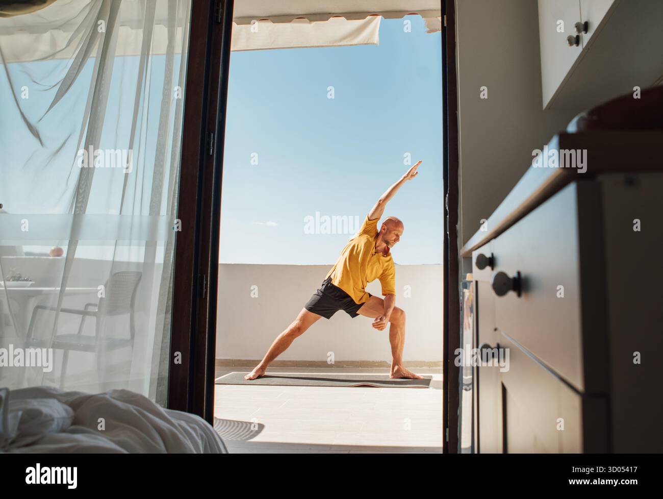 Middle-aged bald man practicing Utthita Parshvakonasana yoga pose on yoga mat on bright balcony during peaceful morning at home. Relaxed lifestyle con Stock Photo
