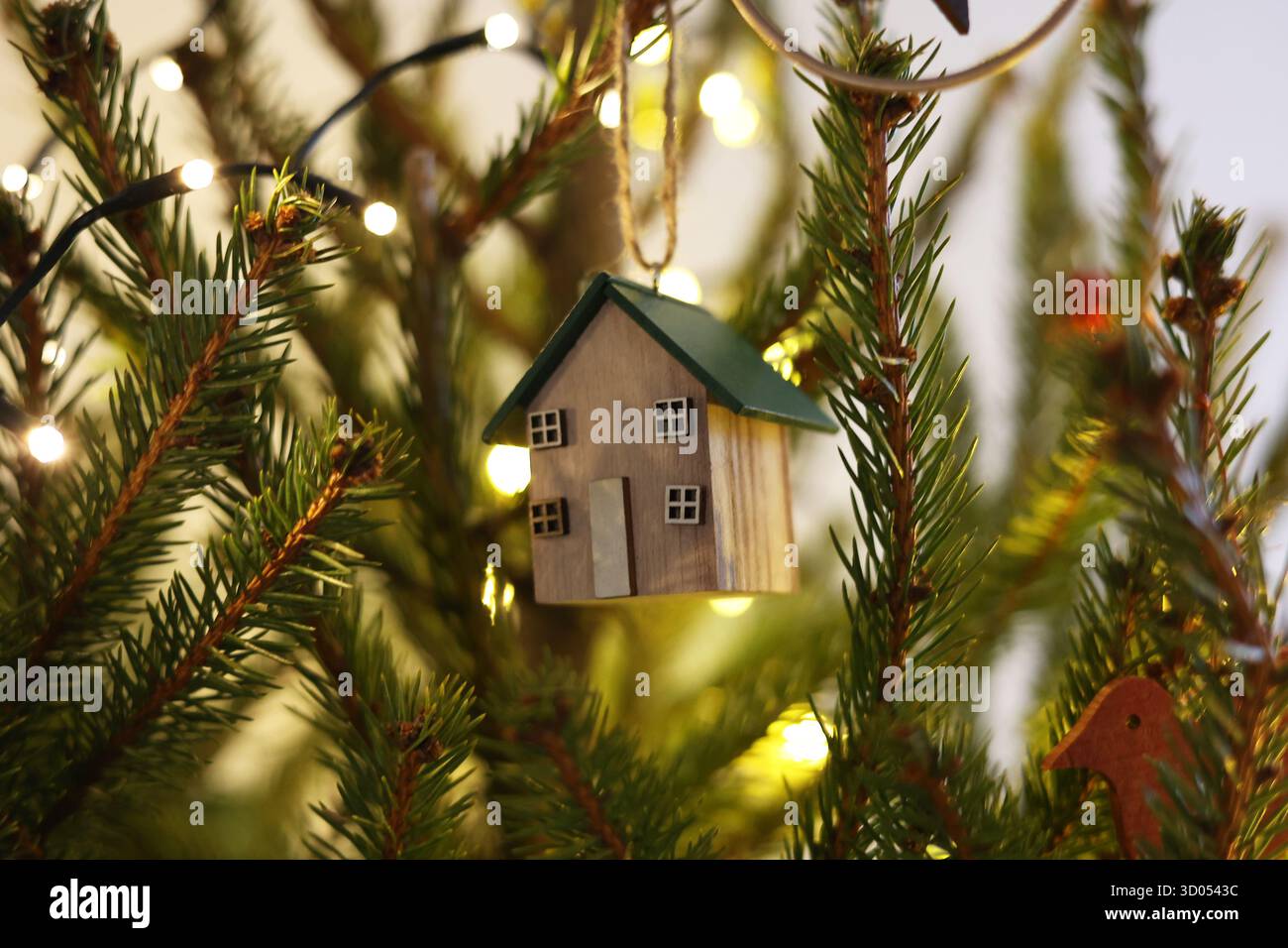 Wooden house ornament hanging on a Christmas tree, surrounded by fir branches and warm fairylights in the background Stock Photo