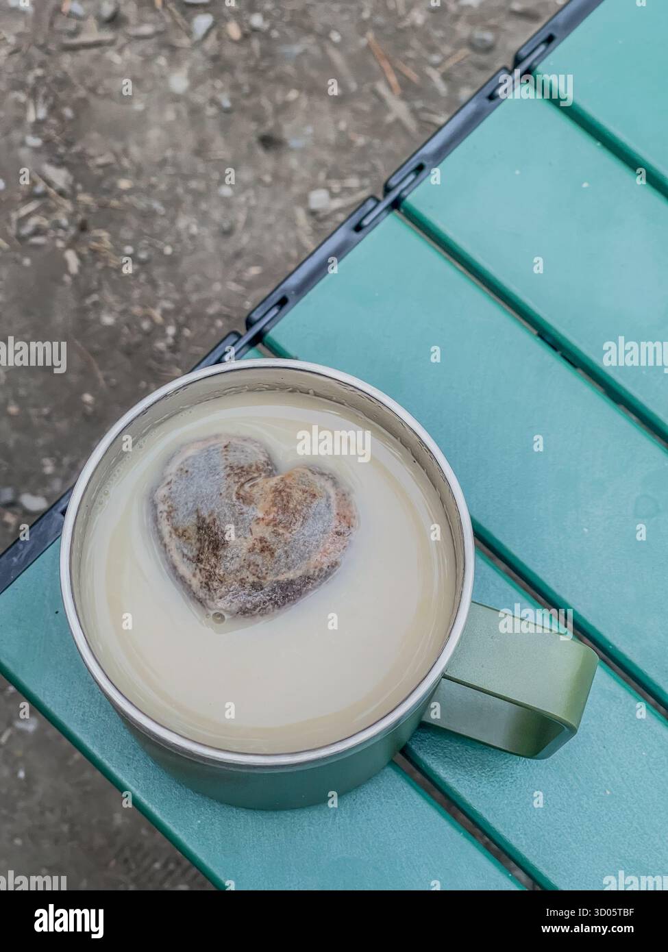 Creamy beverage in a metal mug sits on a green outdoor table. A heart-shaped tea bag floats, adding a whimsical touch to camping - Smartphone Captured Stock Image