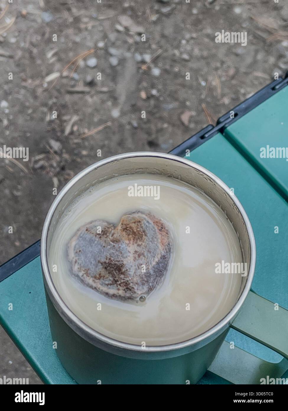 Creamy beverage in a metal mug sits on a green outdoor table. A heart-shaped tea bag floats, adding a whimsical touch to camping - Smartphone Captured Stock Image