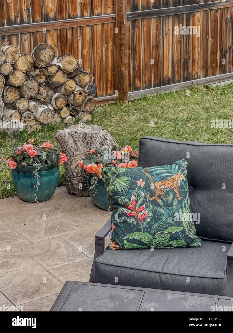 An inviting backyard setup featuring a stacked wood pile, tree stump, flowering pots, and a tropical-patterned cushion on a dark gray outdoor chair - Smartphone Captured Stock Image