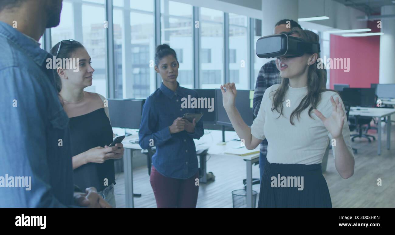 Gesturing woman using VR headset in windowed office, colleagues holding devices, copy space Stock Photo