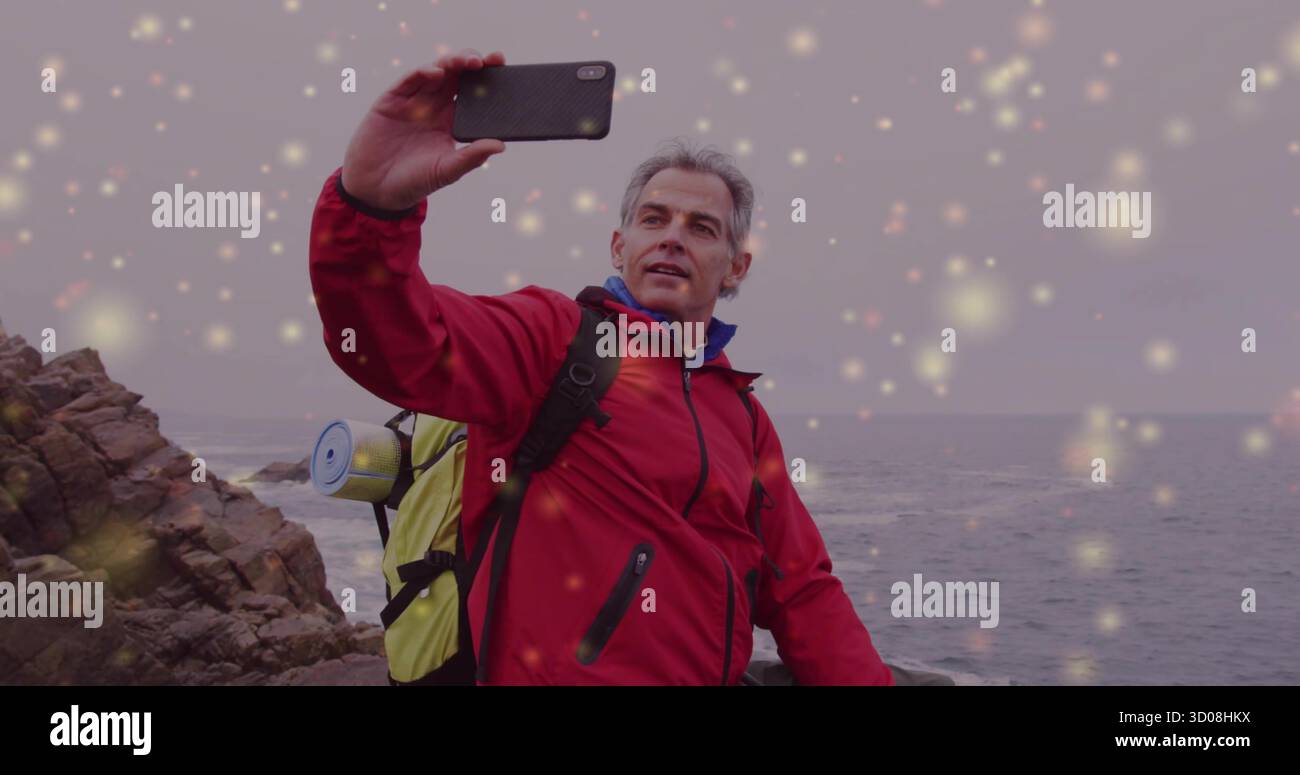 Hiker taking selfie in red jacket on rocky coast, with backpack water bottle smartphone sparkles Stock Photo