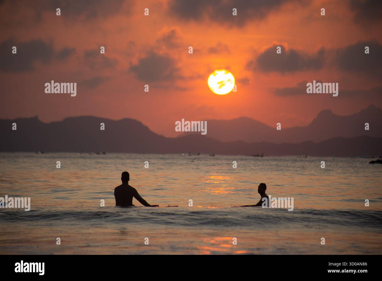 Two people relax in the sea during a stunning sunset at Selong Belanak Beach, Central Lombok, Indonesia, with golden sunlight reflecting on calm water Stock Photo