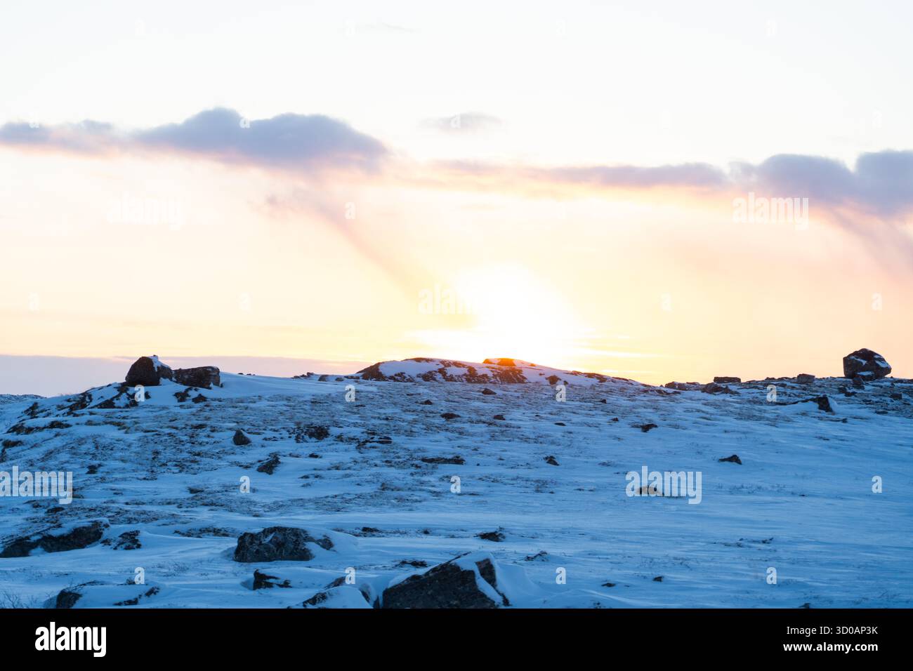 Winter landscape featuring a sun setting behind a snow covered rocky hill with soft pastel clouds in the sky Stock Photo