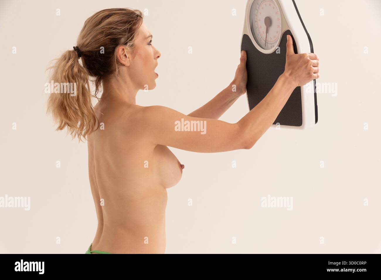 Salisbury Wiltshire England UK. 17.08.2023.  Topless woman  showing signs of anger towards the bathroom scales. Studio shoot. - Stock Image