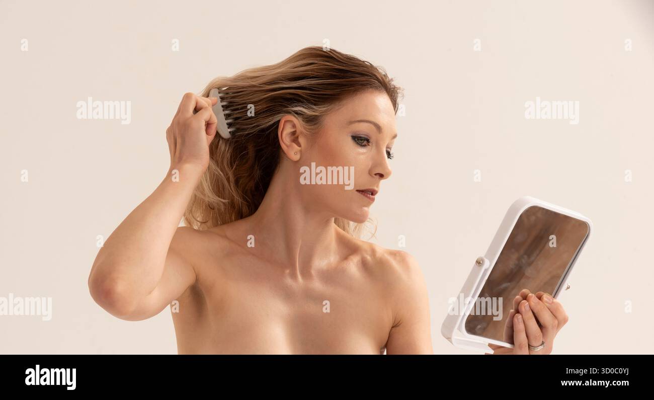 Salisbury Wiltshire England UK. 17.08.2023.  Topless woman using  a mirror and  a comb to style her hair. Studio shoot. - Stock Image