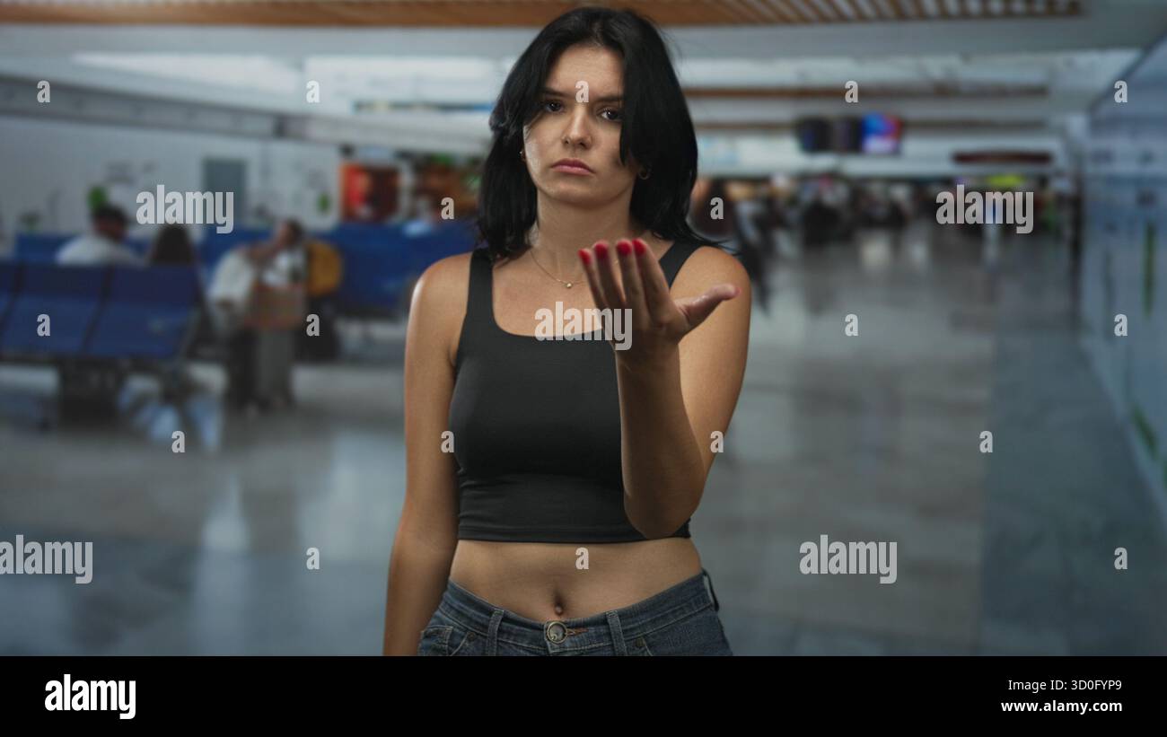 Young hispanic woman beckons with open hand at crowded airport terminal; delayed flight impatience. Stock Photo