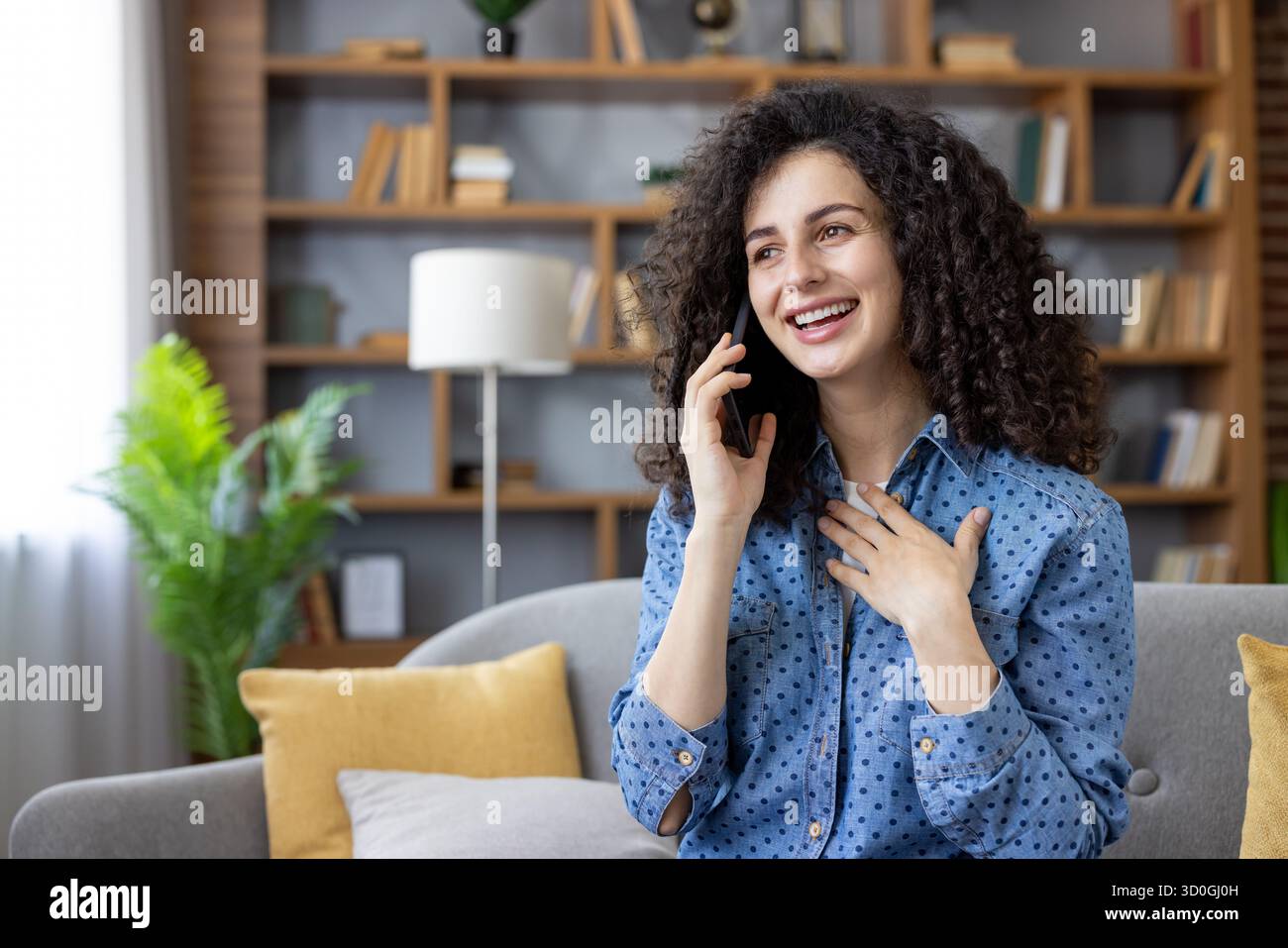 Young woman with curly hair smiling and talking on a mobile phone, relaxed on a cozy living room couch at home, sharing joyful conversation and feeling contentment Stock Photo