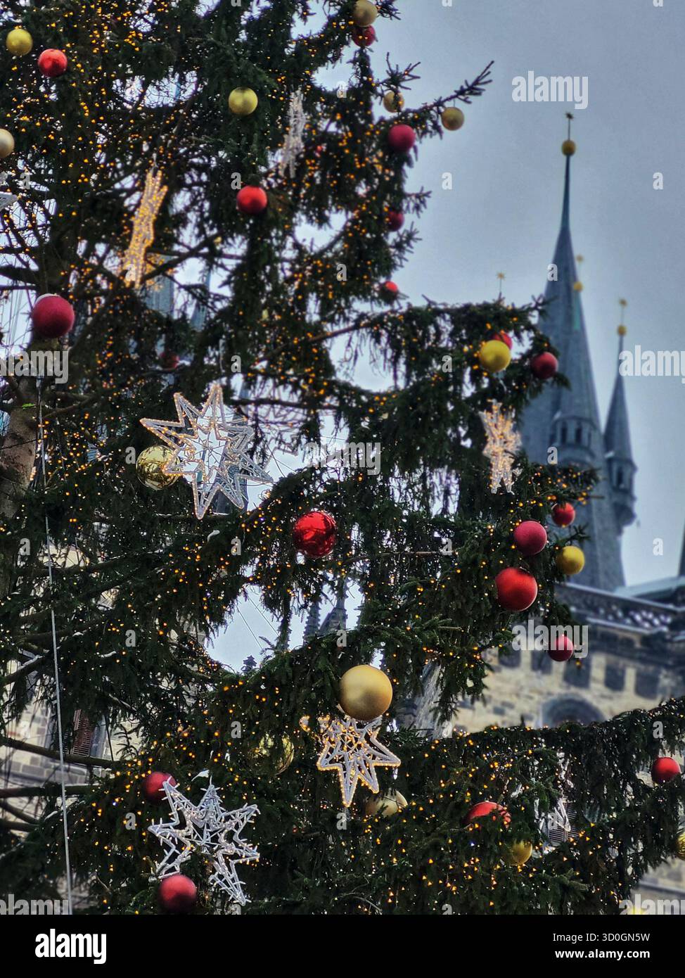 Festive Christmas tree decorated with golden and red ornaments and twinkling lights in front of old gothic towers on a cloudy winter day in a European - Smartphone Captured Stock Image