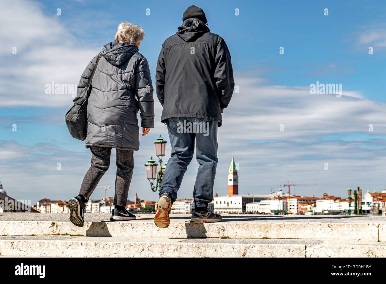 Two people walk across a canal bridge in the Castello district of Venice, with St Mark's Campanile or bell tower in the distance,Venice ,Italy Stock Photo