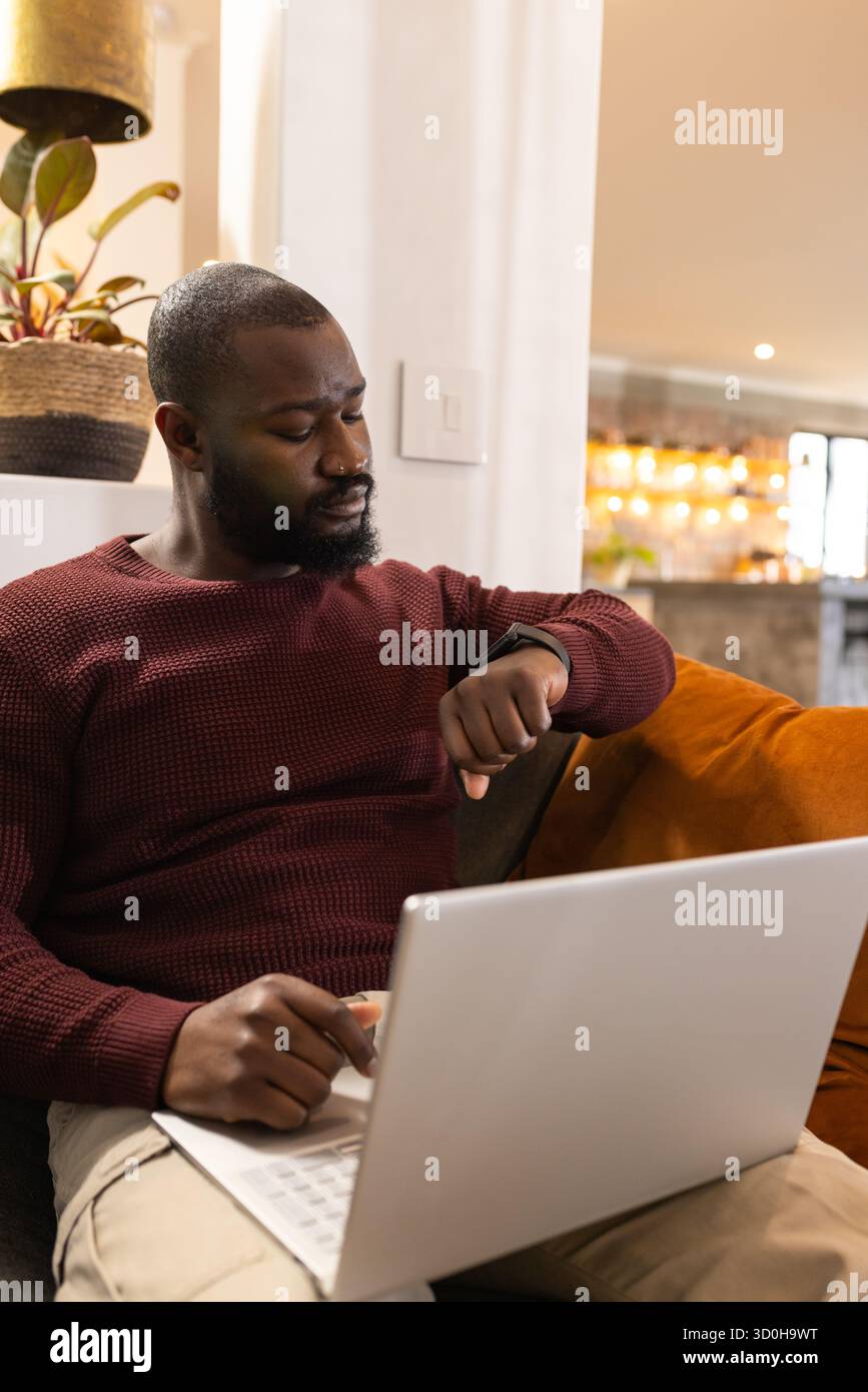 African American man sitting on sofa using laptop and glancing at ...