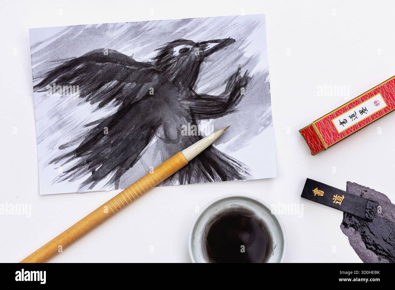 flatlay of a black ink painting of a crow created using japanese ink. Image also features a brush, ink stick, grinding slate, and cereamic dish. - Stock Image