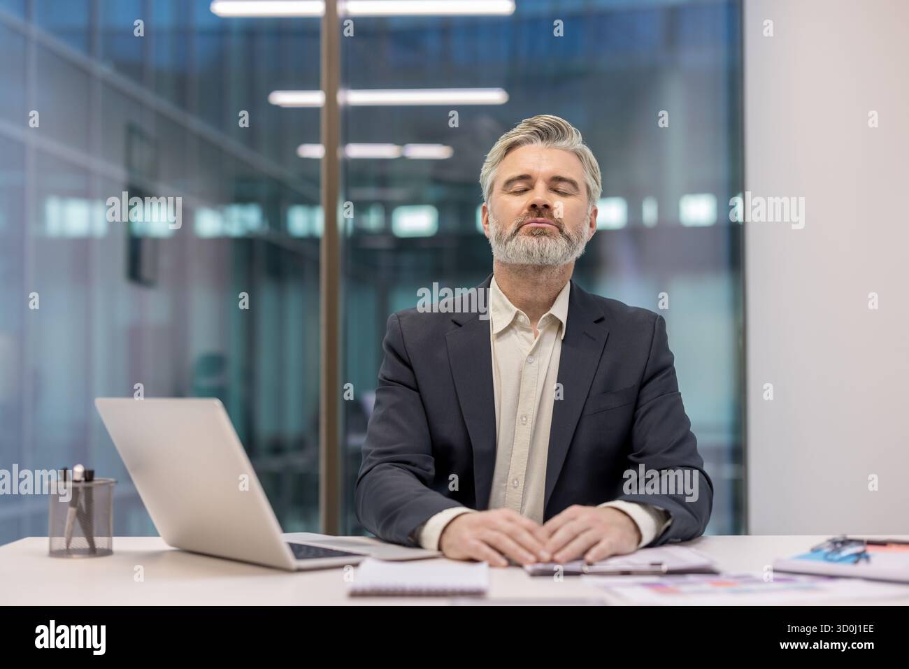 Mature businessman with eyes closed and hands clasped, practicing mindfulness and deep breathing at a modern office desk to relieve stress and restore calm during the workday Stock Photo