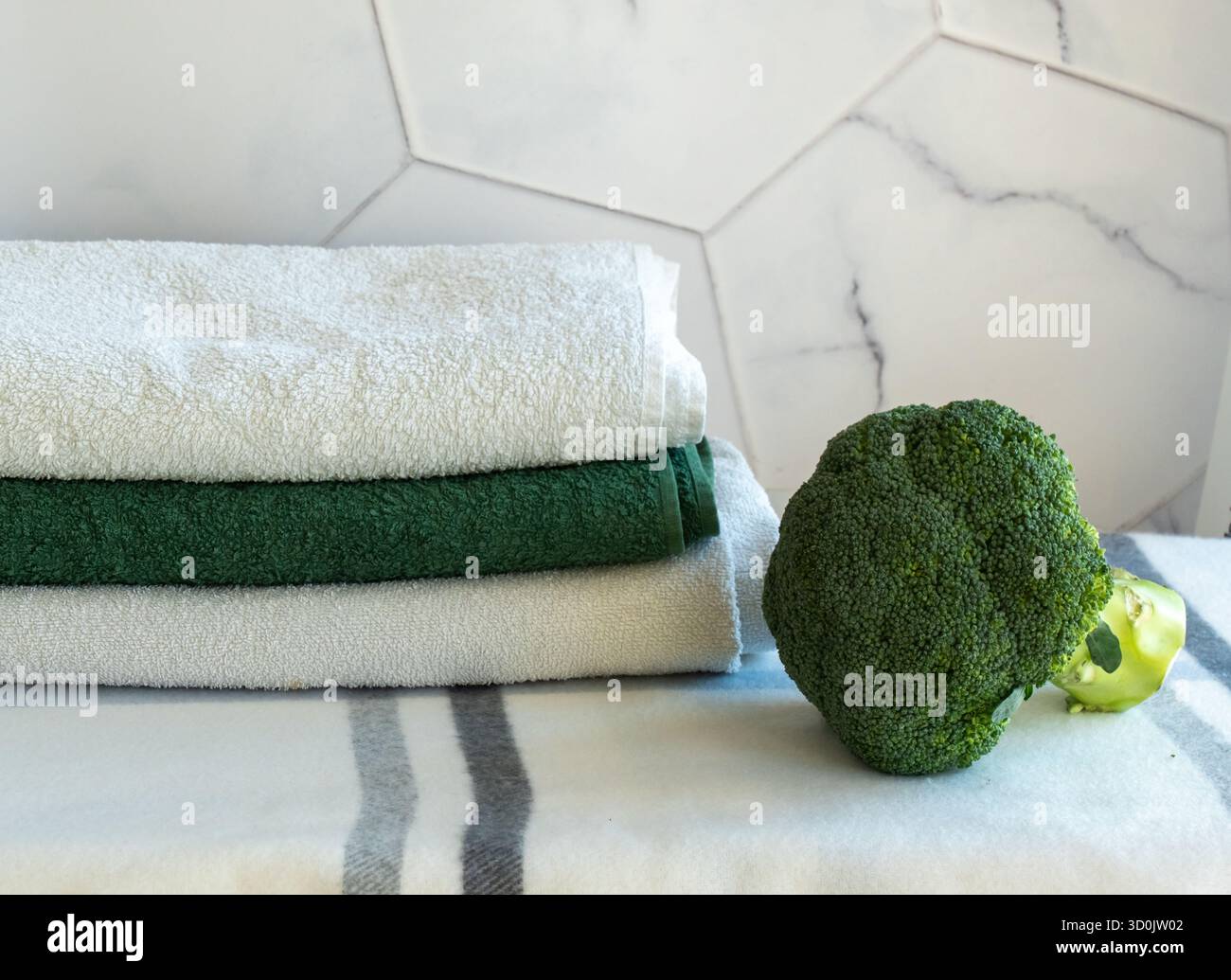 Fresh green broccoli vegetable on marble kitchen counter near stack of clean towels with hexagonal tile background. Healthy cooking and kitchen hygien Stock Photo