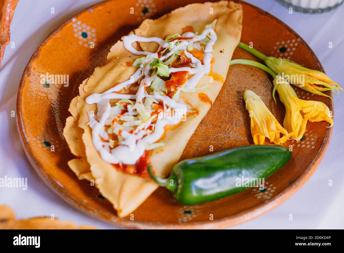 Primer plano de un antojito mexicano relleno, cubierto con salsa y crema, servido en plato de barro con chile y flor de calabaza cruda. Stock Photo