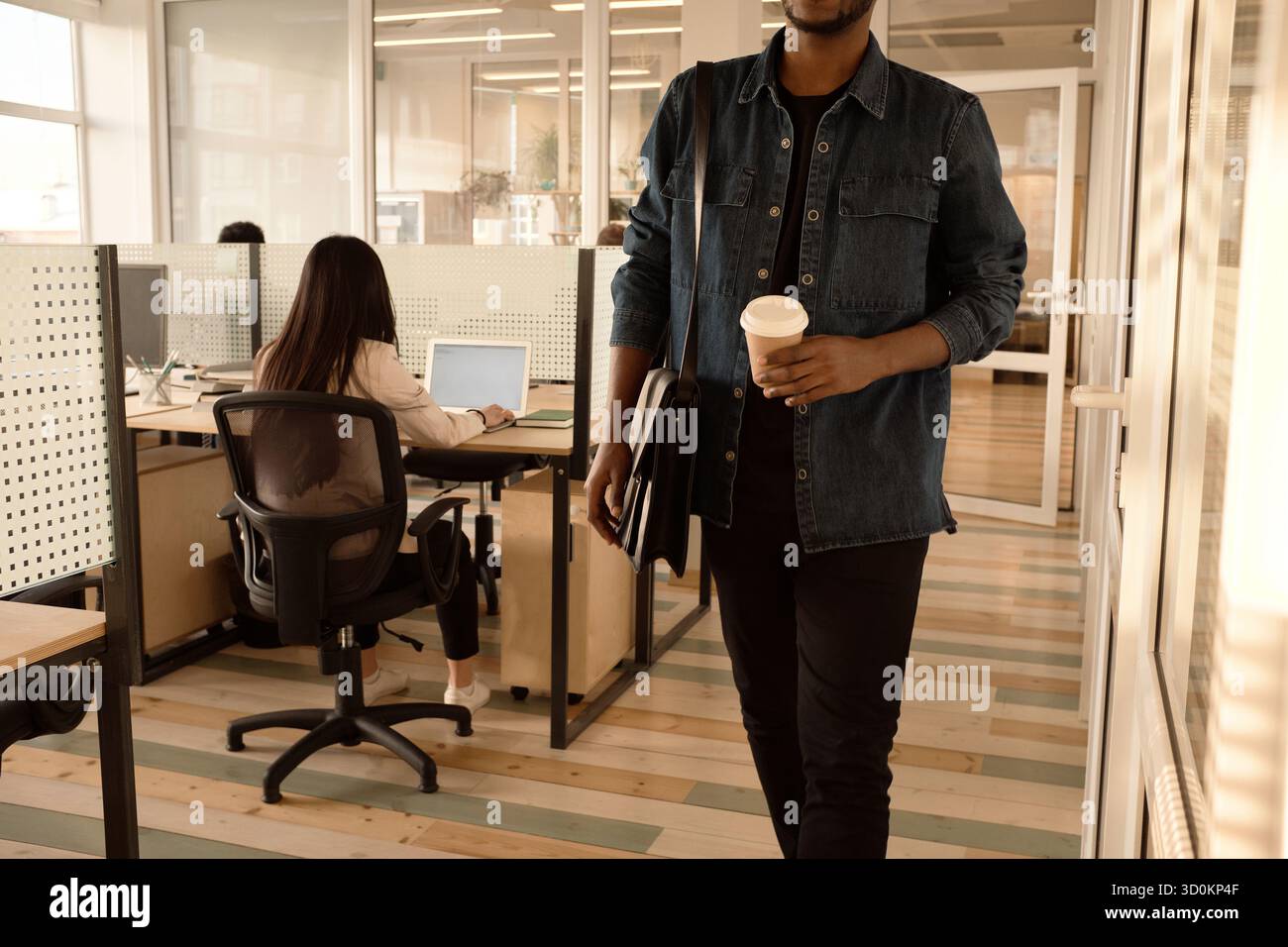 End of working day. Unrecognizable black man leaving office with coffee to go, unrecognizable woman working on laptop behind Stock Photo